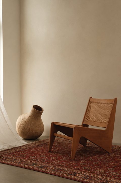 A cane and teak chair on a deep red Persian rug, next to a round woven vessel in a bare, warm-lit room.