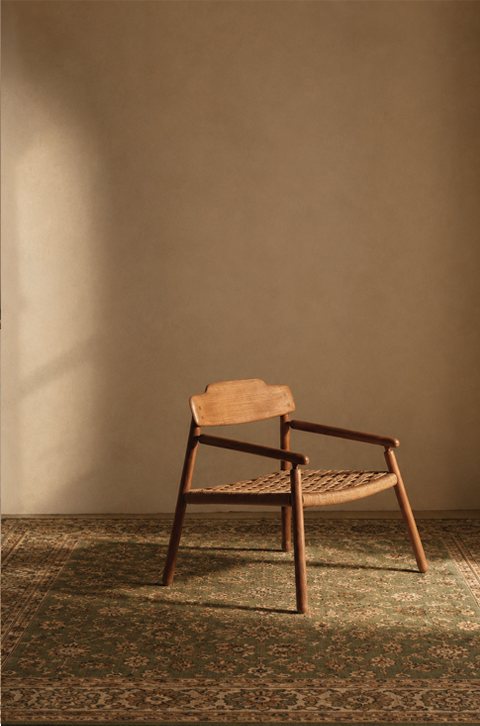 A slender wooden chair standing on a dark green patterned rug against a bare curved plaster wall.
